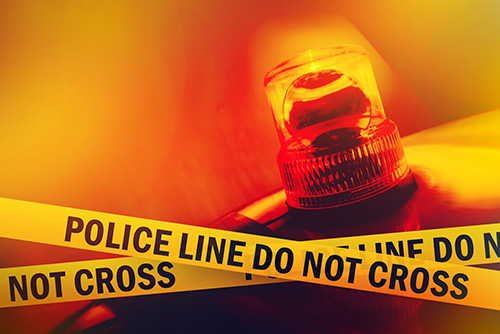 Close-up of police siren light behind yellow 'Police Line Do Not Cross' tape, with a red and orange background suggesting emergency or crime scene.
