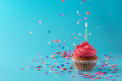 bright pink cupcake with a candle on a blue background with confetti around it.