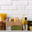 Pantry food staples including pasta, oil, beans, popcorn on a white counter against subway tile in a kitchen setting.