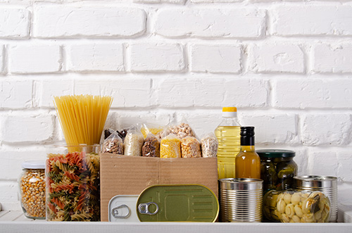 Pantry food staples including pasta, oil, beans, popcorn on a white counter against subway tile in a kitchen setting.
